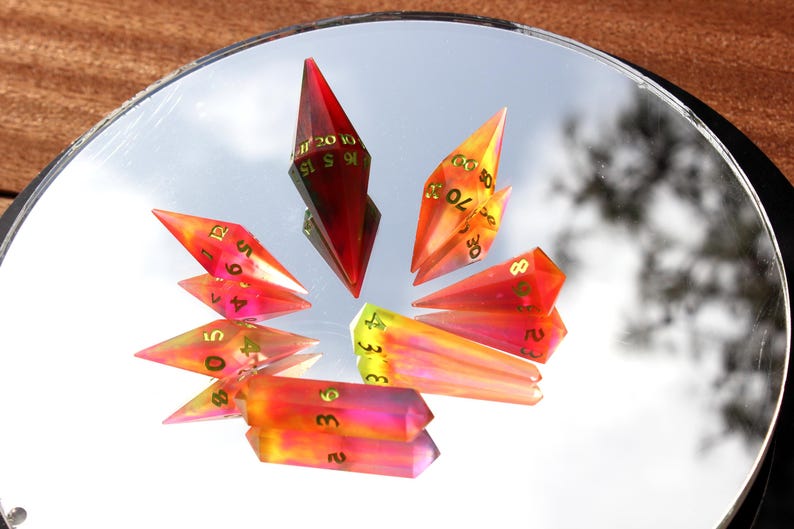May include: A collection of colorful, geometric dice with gold numbering, reflecting on a mirrored surface. The dice are primarily red, orange, and yellow, with a few in shades of pink and purple. The dice are arranged in a circular pattern.