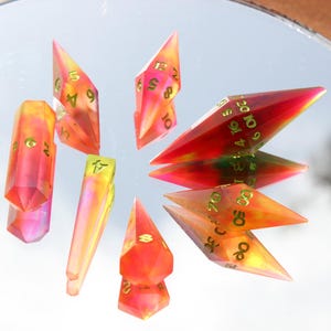 May include: A collection of colorful, translucent polyhedral dice with gold-colored numbers. The dice are in shades of red, orange, and yellow, and are arranged on a reflective surface. The dice are used for tabletop games.
