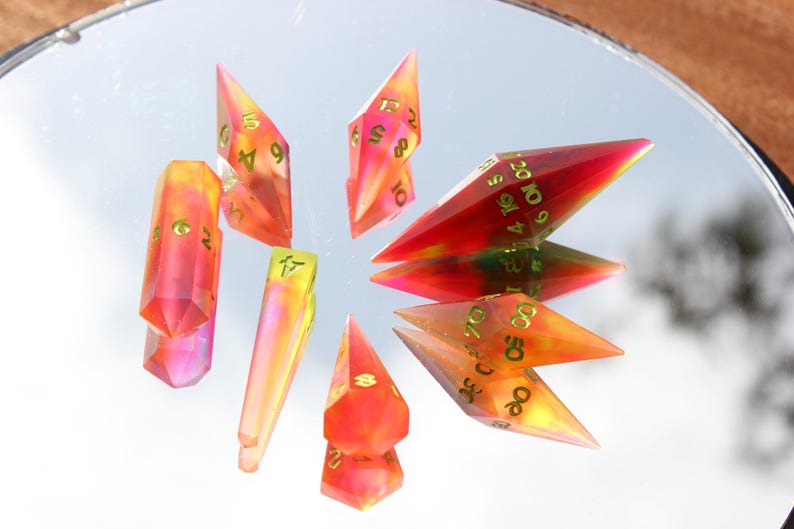May include: A collection of colorful, translucent polyhedral dice with gold-colored numbers. The dice are in shades of red, orange, and yellow, and are arranged on a reflective surface. The dice are used for tabletop games.