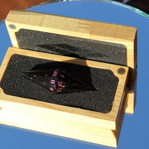 Op de afbeelding: Een zwarte, veelzijdige dobbelsteen met roze cijfers, gepresenteerd in een open houten kist met een schuimrubberen inzetstuk. De dobbelsteen heeft een unieke vorm en de doos is gemaakt van lichtgekleurd hout.