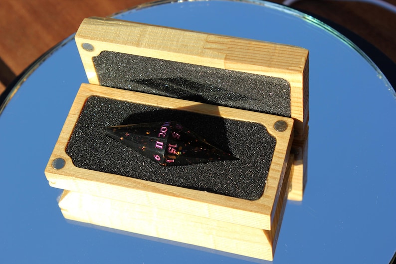 Op de afbeelding: Een zwarte, veelzijdige dobbelsteen met roze cijfers, gepresenteerd in een open houten kist met een schuimrubberen inzetstuk. De dobbelsteen heeft een unieke vorm en de doos is gemaakt van lichtgekleurd hout.