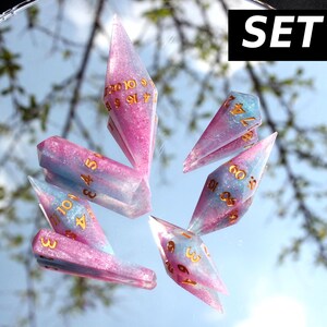 Puede incluir: Un juego de seis dados poli&eacute;dricos con un acabado de purpurina rosa y azul y n&uacute;meros dorados. Los dados est&aacute;n dispuestos sobre una superficie reflectante, y una etiqueta negra en la esquina superior derecha muestra el texto blanco SET #3.