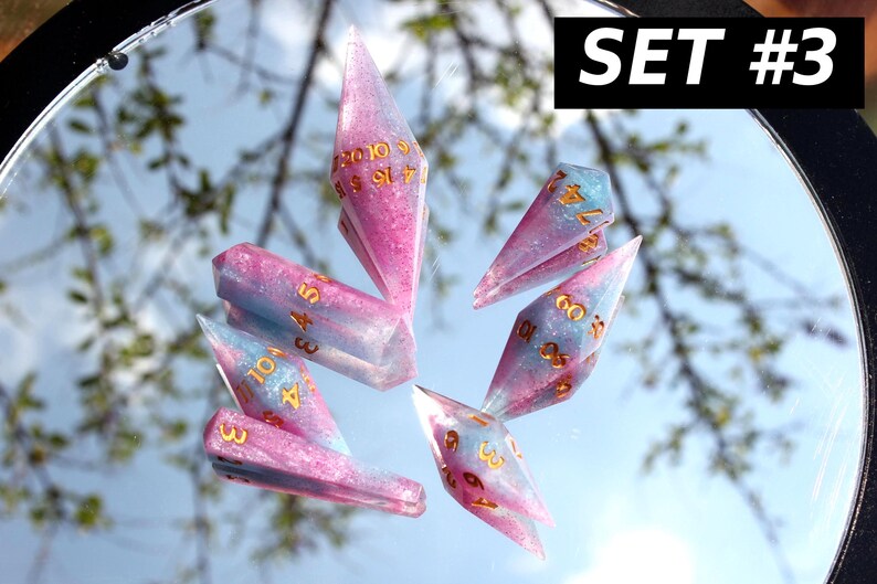 Puede incluir: Un juego de seis dados poli&eacute;dricos con un acabado de purpurina rosa y azul y n&uacute;meros dorados. Los dados est&aacute;n dispuestos sobre una superficie reflectante, y una etiqueta negra en la esquina superior derecha muestra el texto blanco SET #3.
