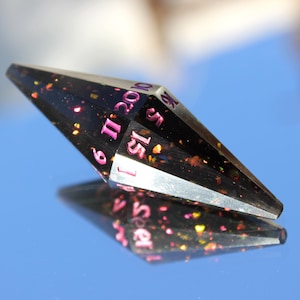 Op de afbeelding: Een zwarte, veelzijdige dobbelsteen met roze cijfers en kleurrijke glitter. De dobbelsteen wordt weerspiegeld op een blauw oppervlak. De cijfers zijn duidelijk zichtbaar, wat duidt op het gebruik ervan in tabletop games.