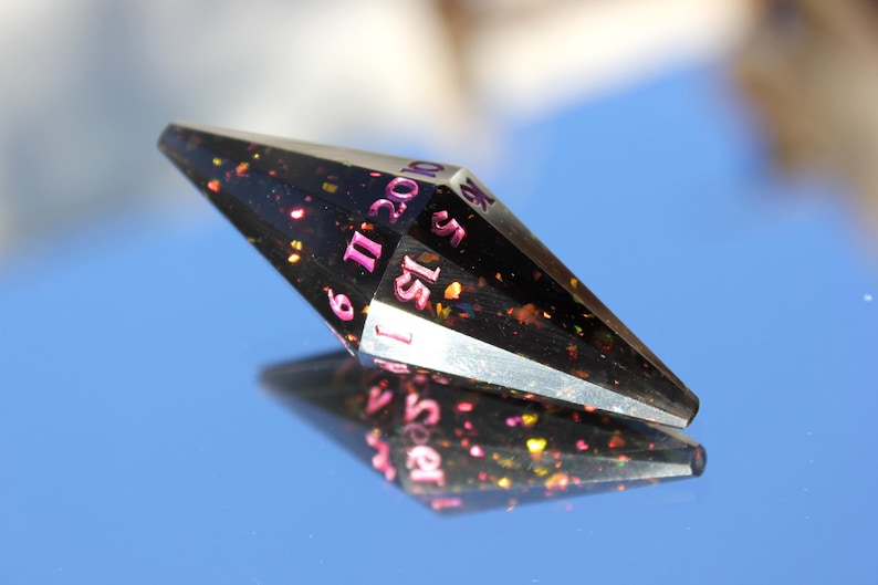 Op de afbeelding: Een zwarte, veelzijdige dobbelsteen met roze cijfers en kleurrijke glitter. De dobbelsteen wordt weerspiegeld op een blauw oppervlak. De cijfers zijn duidelijk zichtbaar, wat duidt op het gebruik ervan in tabletop games.