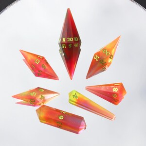 May include: A set of seven polyhedral dice in various shapes, arranged in a circular pattern. The dice are primarily red and orange with gold numbering. The dice are made of a translucent material, and the numbers are clearly visible.