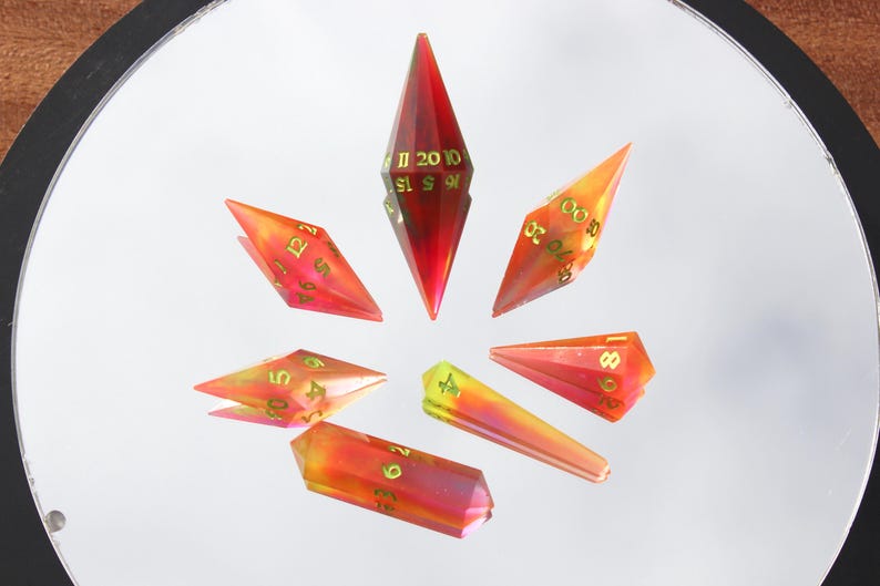 May include: A set of seven polyhedral dice in various shapes, arranged in a circular pattern. The dice are primarily red and orange with gold numbering. The dice are made of a translucent material, and the numbers are clearly visible.