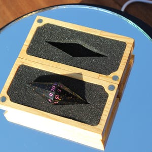 Op de afbeelding: Een houten doos met een zwart schuimrubberen inzetstuk bevat een zwarte en transparante veelvlakkige dobbelsteen met roze cijfers. De doos is open en onthult de dobbelsteen. De doos staat op een reflecterend oppervlak.