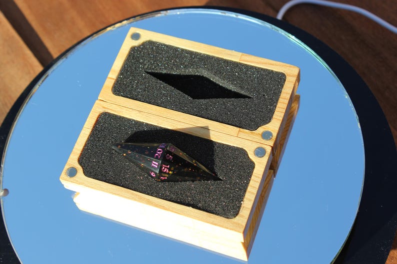 Op de afbeelding: Een houten doos met een zwart schuimrubberen inzetstuk bevat een zwarte en transparante veelvlakkige dobbelsteen met roze cijfers. De doos is open en onthult de dobbelsteen. De doos staat op een reflecterend oppervlak.
