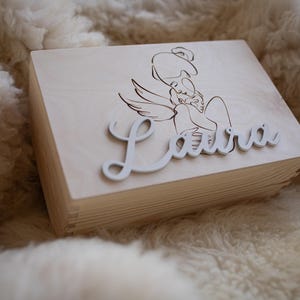 Könnte beinhalten: Eine Holzkiste mit hellbrauner Oberfläche. Die Kiste hat einen Deckel mit einem lasergravierten Bild eines Engels, der ein Baby hält. Der Name "Laura" ist in weißen Buchstaben auf dem Deckel geschrieben.