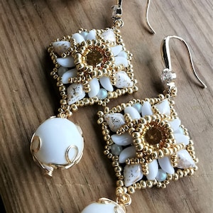 Puede incluir: Un par de pendientes de cuentas doradas y blancas con un colgante de piedra blanca. Los pendientes están hechos con cuentas pequeñas y redondas y tienen una forma cuadrada. El colgante es una piedra blanca que está rodeada por un alambre dorado.