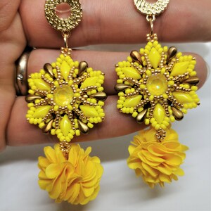 Może przedstawiać: Para żółtych kolczyków z koralików z kwiatem. Kolczyki są wykonane z małych żółtych koralików i złotych akcentów. Kwiatowy element jest wykonany z żółtej tkaniny.