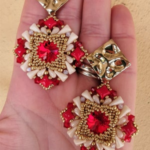 May include: A pair of red and gold earrings with a square design. The earrings feature a large red crystal in the center, surrounded by smaller red and white beads. The earrings are attached to a gold square pendant.