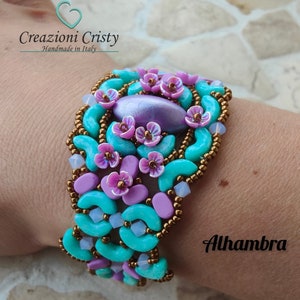 May include: A turquoise and purple beaded bracelet with a large iridescent purple bead in the centre. The bracelet is decorated with small purple flowers and the text "Alhambra" is visible on the wrist.
