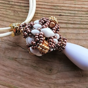 Può includere: Una collana di perline bianche e marroni con un ciondolo bianco a forma di lacrima. La collana è realizzata con perline piccole e ha una chiusura dorata.