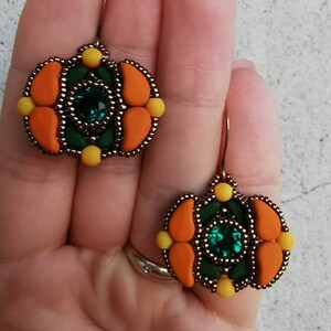 Può includere: Un paio di orecchini fatti a mano con un design a forma di zucca. Gli orecchini hanno un centro verde scuro con una gemma quadrata color smeraldo, circondata da accenti arancioni e gialli. I bordi sono delineati da piccole perline scure.