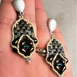 May include: A pair of silver and brown beaded earrings with a woven design and teal gemstones. The earrings are hanging from a person's ear.