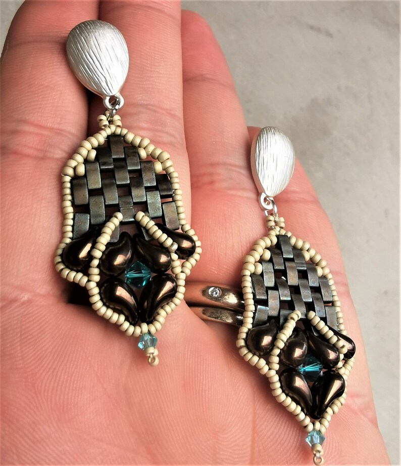 May include: A pair of silver and brown beaded earrings with a woven design and teal gemstones. The earrings are hanging from a person's ear.