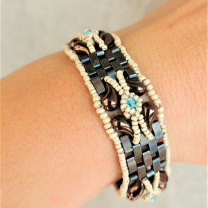 May include: A brown and white beaded bracelet with a blue gemstone accent. The bracelet is made with small, square beads and features a woven pattern.