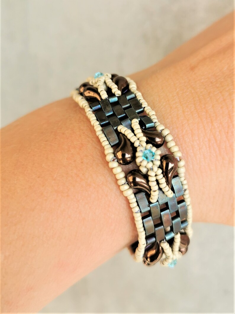 May include: A brown and white beaded bracelet with a blue gemstone accent. The bracelet is made with small, square beads and features a woven pattern.