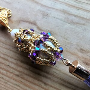 May include: A gold chain necklace with a purple and gold beaded pendant featuring a white pearl. The pendant has a tassel attached to the bottom.