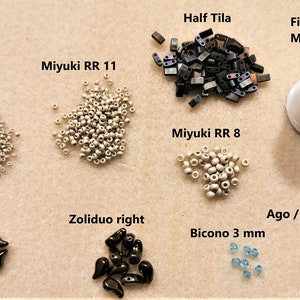 May include: Assortment of beads and beading supplies for jewellery making, including white seed beads, black teardrop beads, blue bicone beads, silver half tila beads, and a spool of white beading thread.