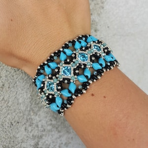 Könnte beinhalten: Ein türkisfarbenes und schwarzes Armband mit silbernen Akzenten. Das Armband hat ein geometrisches Muster mit quadratischen türkisfarbenen Perlen.