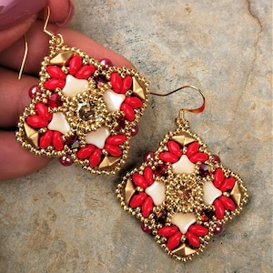 Può includere: Un paio di orecchini a forma quadrata con perline dorate, rosse e bianche, con un design geometrico. Gli orecchini presentano un cristallo centrale.