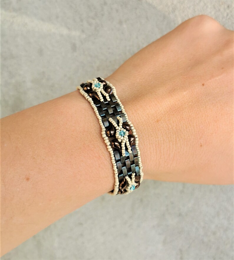 May include: A brown and white beaded bracelet with blue accents. The bracelet is woven with small beads and features a geometric pattern.