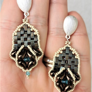 Peut inclure: Une paire de boucles d'oreilles pendantes avec un haut en forme de larme argenté, un motif tissé de perles gris foncé et brunes, et un groupe de perles brunes en forme de larme avec un cristal bleu au centre.