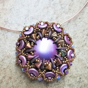 Könnte beinhalten: Eine violette und goldene Perlenkette mit einem großen, schillernden violetten Anhänger. Der Anhänger ist von kleineren Perlen in Braun-, Gold- und Violetttönen umgeben.