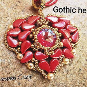 Può includere: Un ciondolo a forma di cuore rosso e oro con un grande cristallo al centro. Il ciondolo è fatto di perline e ha un design gotico. Il testo "Gothic heart" è scritto sopra il ciondolo.