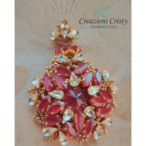 May include: A large, ornate pendant featuring a cluster of red and clear faceted crystals set in gold tone metal. The pendant is designed in a teardrop shape and has a delicate, intricate design.
