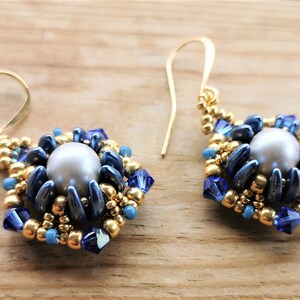 May include: A pair of gold dangle earrings with a blue and gold beaded design. The earrings feature a large pearl-like bead in the center of each earring.