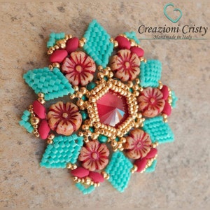 Könnte beinhalten: Eine türkisfarbene und goldene Perlenblume mit einem roten Kristall in der Mitte. Die Blume ist von kleineren roten und weißen Perlen umgeben. Der Text "Creazioni Cristy Handmade in Italy" ist in der oberen rechten Ecke des Bildes zu sehen.