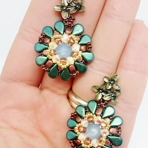 Op de afbeelding: Een paar groene en bruine kralen oorbellen met een bloemmotief. De oorbellen hebben bovenaan een cluster van kleine metalen bloemen en een grotere kralenbloem met een helder kristal in het midden.