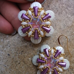 May include: A pair of purple and white beaded earrings with gold accents and a crystal in the center. The earrings are shaped like a flower.