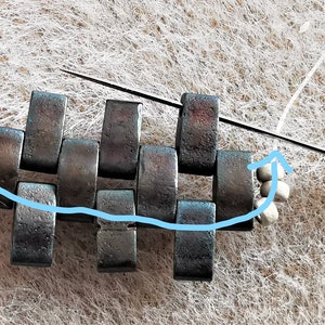 May include: A close-up of a metal chainmail bracelet being made with silver beads and a needle and thread. The thread is being threaded through the metal links of the bracelet.