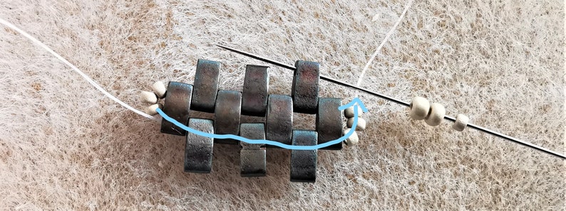 May include: A close-up of a metal chainmail bracelet being made with silver beads and a needle and thread. The thread is being threaded through the metal links of the bracelet.