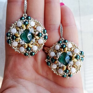 Può includere: Un paio di orecchini quadrati di colore verde e oro con perline bianche e una grande pietra preziosa verde al centro. Gli orecchini hanno un design delicato.