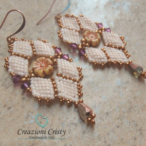May include: A pair of white and gold beaded earrings with a flower design and purple accents. The earrings have a teardrop-shaped pendant with a iridescent finish.