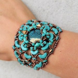 Könnte beinhalten: Ein türkisfarbenes Perlenarmband mit einem großen Mittelstein und kupferfarbenen Akzenten. Das Armband hat halbmondförmige Perlen und kleine runde Perlen, die ein strukturiertes Design ergeben. Das Armband ist etwa 5 cm breit.