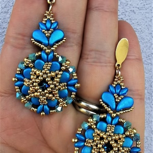 Può includere: Un paio di orecchini pendenti con un designo di perline blu e dorate. Gli orecchini presentano un centro rotondo di perline con una perlina blu più piccola al centro. Gli orecchini sono sospesi da un gancio dorato.