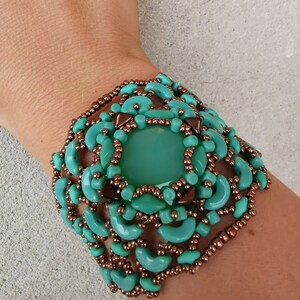 Könnte beinhalten: Ein türkisfarbenes und kupferfarbenes Armband mit einem großen türkisfarbenen Cabochon in der Mitte. Das Armband ist aus Perlen gefertigt und hat ein zartes, filigranes Design.
