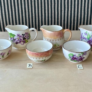 Puede incluir: Cinco tazas de té vintage y platillos con diseños florales. Las tazas son blancas con patrones florales morados y verdes. Los platillos son blancos con ribetes dorados. Las tazas están etiquetadas con A, B y C.