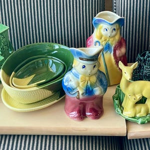 Pode incluir: Uma coleção de estatuetas e vasos de cerâmica vintage. As estatuetas incluem uma mulher com um vestido verde, um menino com um traje azul e vermelho, e um veado com um filhote. Os vasos incluem um moinho de vento, um poço e um par de tigelas aninhadas.