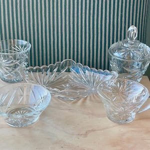 Könnte beinhalten: Eine Sammlung klarer Servierteile aus geschliffenem Glas. Das Set umfasst einen Becher, eine kleine Schale, eine Tasse mit Henkel, eine Schale mit Deckel und eine lange, gewellte Schale. Das Glas hat ein Stern- und geometrisches Muster.