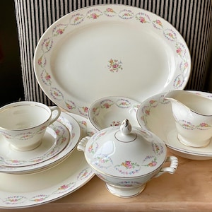 May include: A vintage porcelain dinner service. The set includes an oval platter, plates, a lidded serving dish, a gravy boat, and teacups. Each piece is white with a floral garland design and gold trim.