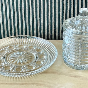 Può includere: Piatto da portata e barattolo con coperchio in vetro trasparente con un design strutturato. Il piatto ha un centro a forma di stella e un bordo a coste. Il barattolo ha un motivo decorativo e un pomello sul coperchio.