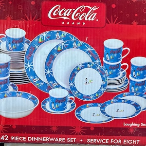 Puede incluir: Un juego de vajilla Coca-Cola de 42 piezas con un diseño de muñeco de nieve riendo. El juego incluye platos, tazones y tazas con un patrón de copo de nieve azul y blanco. La caja dice "Servicio para ocho personas".